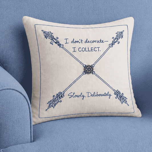 Decorative pillow with blue “I don’t decorate — I collect” lettering and crossed ornamental arrows displayed on a blue upholstered chair.