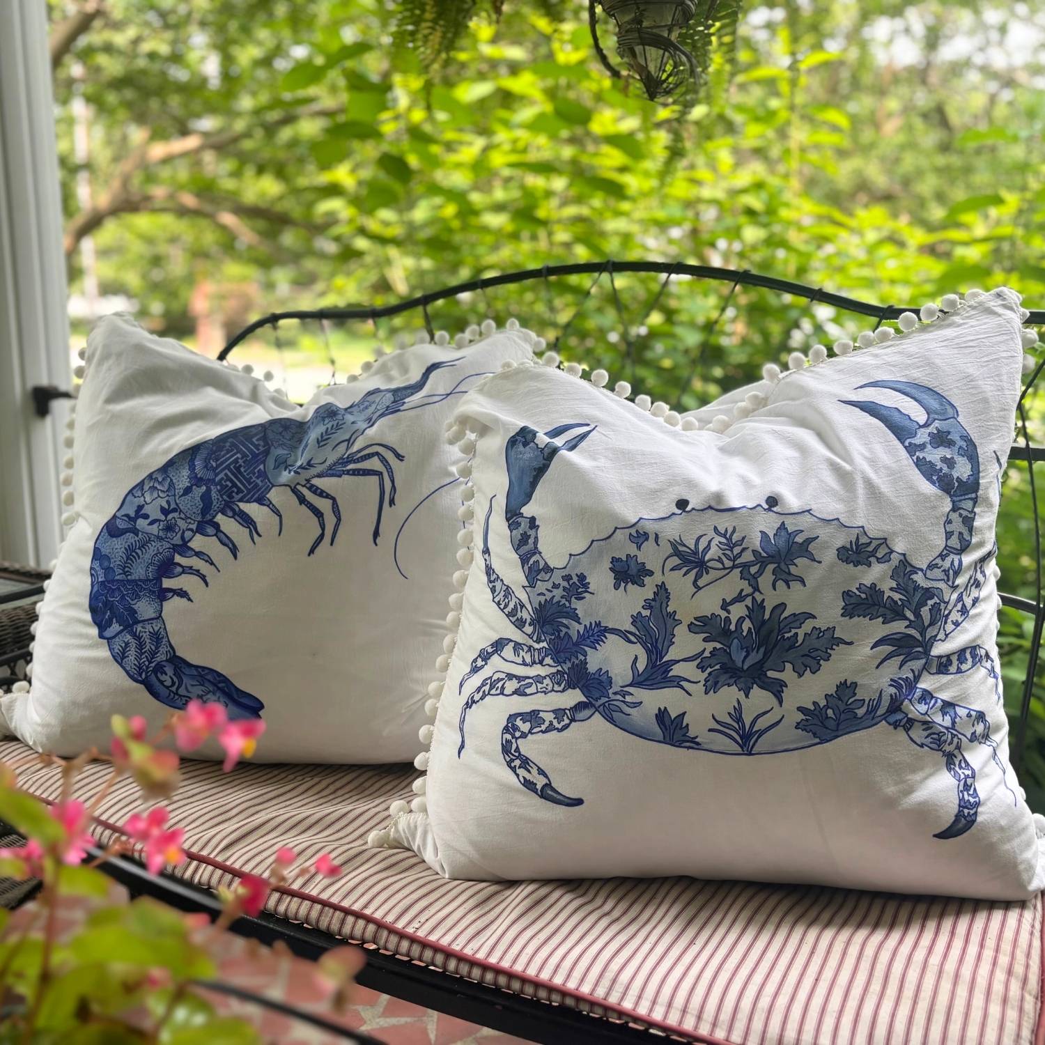 Two hand-painted chinoiserie-style crab and prawn pillows with white pom pom trim, styled on a bench.