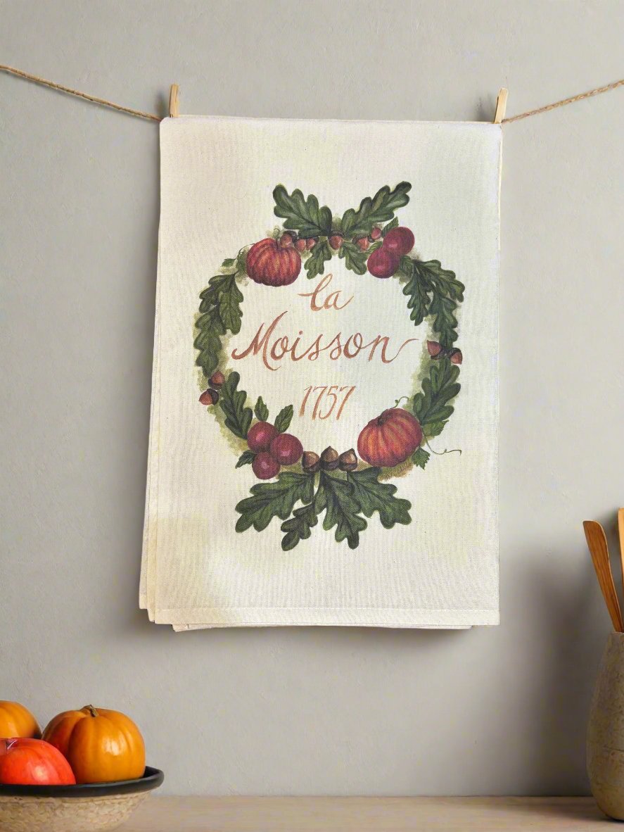 Photo of a white cotton tea towel with a fall harvest wreath design featuring pumpkins, cranberries, acorns, and green oak leaves. It is hanging on twine with clothes pins on a kitchen wall, and small decorative pumpkins are in a bowl on the countertop. 